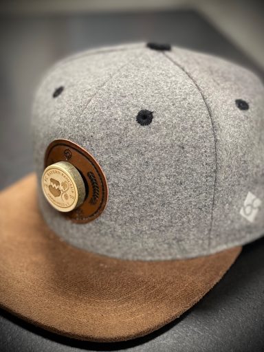 Grauer Wollhut mit brachendem Schirm und metallenem Logo.