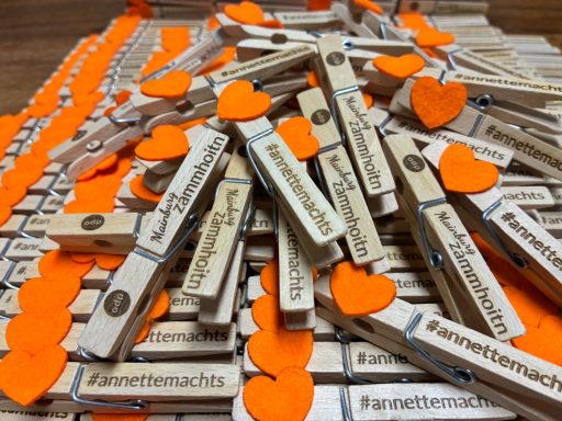 ÖDP Mainburg Bunte Wäscheklammern mit Herzen, angeordnet auf einem orangefarbenen Untergrund.