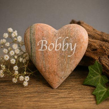 Herzförmiger Stein mit der Aufschrift "Bobby" und kleinen weißen Blüten daneben.