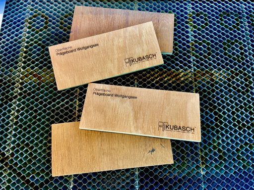 KUBASCH Fenster & Türen GmbH Vier Holzplatten mit Gravuren, gestapelt auf einem Gitteruntergrund.