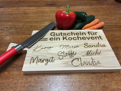 Ein Holzbrett mit Gravur von Namen und einem Gutschein für ein Kochevent, umgeben von Gemüse.